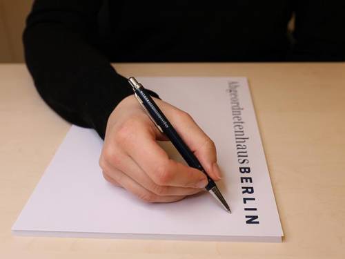 Man sieht eine Hand, die einen Stift hält und auf einem Notizblock zu schreiben beginnt. Auf dem Notizblock steht gedruckt "Abgeordnetenhaus Berlin"